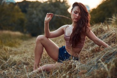 Gün batımında samanlıkta poz veren güzel seksi genç bir kadın. Kot şort ve bej bluz giymiş, doğada bir yaşam tarzı var. Romantik sonbahar havası, altın saat