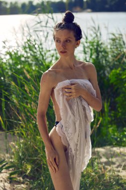 Sazlıkta poz veren güzel model. Baştan çıkarıcı kadın göğsünü ve gövdesini temiz beyaz bir havluyla kaplıyor, suyun yanında dinleniyor.