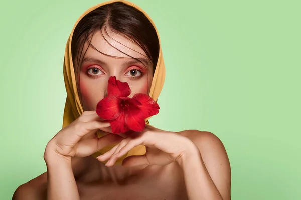 一个漂亮的女人 手上拿着红花 靠近脸庞 她的头披着时髦的头巾 赤裸的肩膀 有着粉红妆容的皮肤模型 绿色背景的护肤和温泉概念 — 图库照片