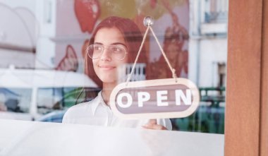Mutlu küçük işletme kızı pencerenin önünde açık tabelayla üstünü değiştirip gülümsüyor.