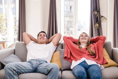 Genç bir çiftin portresi evdeki kanepede otururken ellerini başının arkasına koyup rahatlatıyor..