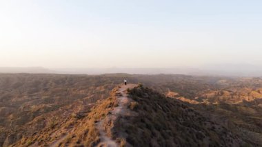 Bir dağ sırtının tepesinde duran kadın, altın bir günbatımında kurak manzaranın güzel ve huzurlu manzarasının tadını çıkarıyor.
