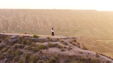Uçurumun kenarında duran tesettürlü bir kadının hava görüntüsü. Altın bir günbatımında engin çöl manzarasını ve kanyonu düşünüyor.