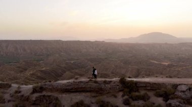 Gün batımında uçsuz bucaksız bir çöl kanyonunda el ele tutuşan ve bir uçurum kenarında yürüyen çiftin görkemli hava manzarası.