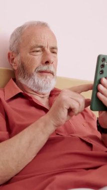 Rahat kıdemli bir adam akıllı telefonuyla internet içeriğine rahatça göz gezdirirken aynı zamanda yatakta rahat bir şekilde uzanıyor, dijital bağlantının ve modern teknolojinin tadını çıkarıyor. Dikey görüntüler