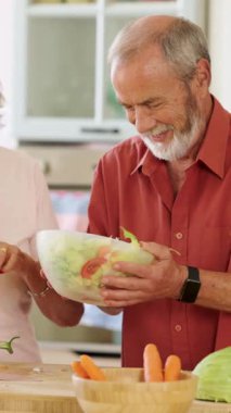 Yaşlı bir adam karısının yardımıyla sağlıklı bir salata hazırlıyor. Mutfaklarındaki bir kaseye baharat ve karışımı katıyor. Dikey görüntüler