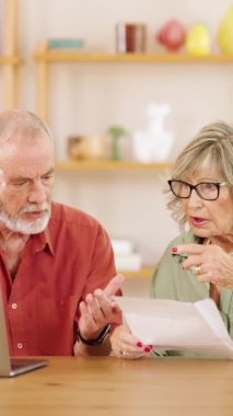 Finansal stres yaşayan üst düzey bir çift, belgeleri dikkatle analiz ediyor ve dizüstü bilgisayar kullanıyor, oturma odası düzenindeki ev giderleriyle ilgili endişelerini paylaşıyor. Dikey görüntüler