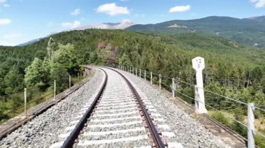 Berrak mavi bir gökyüzünün altında yemyeşil bir dağ ormanında kıvrılan demiryolu raylarının manzarası