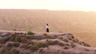 Havadan insansız hava aracı tesettürlü Müslüman bir kadından geri çekiliyor. Uçurumun kenarında duruyor. Uçsuz bucaksız, çorak bir kanyona bakıyor.