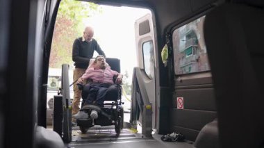 Fiziksel engelli bir adam tekerlekli sandalye kullanarak özel donanımlı bir minibüse biniyor. Bakıcının yardımıyla, ulaşılabilir seyahat imkanı sağlıyor.