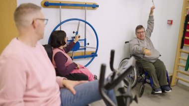 Uzman bir spor salonunda egzersiz yapan fiziksel engelli bir grup kararlı insan. Tekerlekli sandalyedeki adam vücudunun üst kısmı için ip çekiyor.