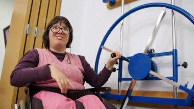 Tekerlekli sandalyede engelli bir kadın, üst vücut rehabilitasyonu için fizik tedavi seansı sırasında özel ekipman kullanıyor.