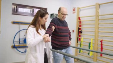 Kendini yaşlı bir adama rehabilitasyon kliniğinde paralel çubuklar kullanarak yürüme ve denge egzersizlerinde yardımcı olmaya adamış bir kadın fizyoterapist.