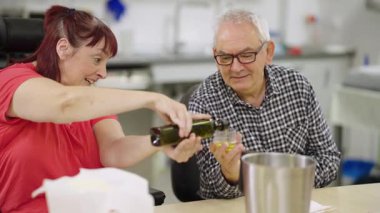 Tekerlekli sandalyede engelli neşeli bir kadın yemek kursunda son sınıf bir adamın tuttuğu şişeden zeytinyağı döküyor.