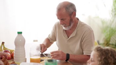 Beyaz sakallı yaşlı bir adam aile yemeği sırasında masada oturuyor. Kahvaltıda bir parça tostun üzerine tereyağı sürüyor.