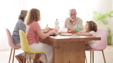 Parlak bir yemek odasında sağlıklı bir kahvaltı yapan üç nesillik mutlu bir aile. Büyükbaba, anne ve çocuk yemek yiyor ve konuşuyor.