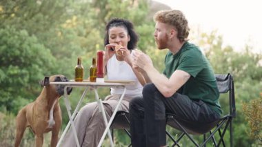 Sosisli yiyen ve parktaki katlanan masada bira içen çok ırklı genç bir çift. Şirin boksör köpekleri onları yerken izliyor.