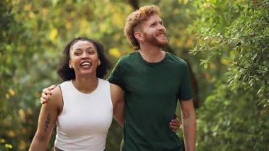 Güneşli bir parkta romantik bir yürüyüş yapan neşeli, ırklar arası genç bir çift. Mutlu adam ve kadın dışarıda kucaklaşırken gülümsüyor ve konuşuyor.