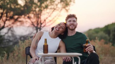 Romantik bir açık hava buluşmasının tadını çıkaran genç bir çift. Mutlu adam ve kadın gün batımında kamp yaparken bira içiyor, gülümsüyor ve konuşuyor.