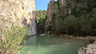 Derin ve kayalık bir kanyondan akan turkuaz bir nehri olan huzurlu bir manzara. Berrak mavi gökyüzünün altındaki sarp kayalıklarda yemyeşil bitki örtüsü büyüyor.