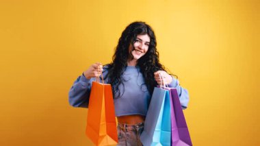 Uzun kıvırcık saçlı, renkli alışveriş torbaları taşıyan ve gülümseyen neşeli genç bir kadın. Sarı arka planda neşeli bir müşterinin stüdyo çekimi.