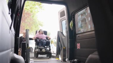 Engelli bir adam tekerlekli sandalyeli bir minibüsü araç olarak kullanıyor. Bir bakıcı ona araç platformunda yardım ediyor.
