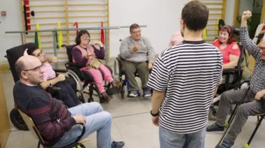 Bir grup engelli erkek ve kadın fizik tedavi seansına katılıyor. Bir antrenör sınıfa yol gösterir, onlara egzersizlerde rehberlik eder.