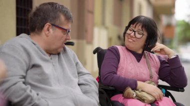 Orta yaşlı bir çift, engelli bir adam ve bir kadın, ikisi de şehir caddesinde tekerlekli sandalyede oturmuş neşeli bir konuşma yapıyorlar.