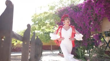 Geleneksel beyaz ve kırmızı flamenko bir elbise giyen zarif bir kadın, çiçeklerle dolu tipik bir İspanyol avlusunda poz verip dans ediyor.