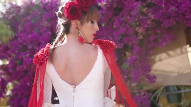 Güzel bir kadın geleneksel beyaz flamenko elbisesi giyiyor ve kırmızı detaylar zarif bir şekilde dans ediyor. Mor çiçeklerle dolu bir bahçede gösteri yapıyor.