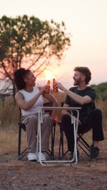 Kamp sandalyelerinde oturan, bira şişelerini kızartan, konuşan ve doğada güzel bir günbatımının tadını çıkaran genç çift.