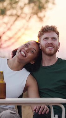 Açık bir kafede oturup birlikte bira içen çok ırklı genç bir çift. Güzel bir kadın partnerinin omzuna yaslanıyor, gülümsüyor.