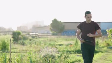 Günbatımında tarlasında yürüyen modern çiftçi. Bitkilerin büyümesini izlemek ve veri toplamak için tablet kullanan genç bir tarım uzmanı.