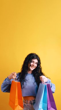 Kıvırcık saçlı neşeli esmer kadın gülümsüyor ve renkli alışveriş torbaları gösteriyor. Parlak sarı bir arka planda alışverişiyle eğlenen neşeli bir müşteri. Dikey