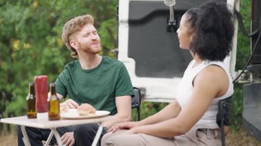 Mutlu milenyum çifti karavanlarının yanındaki masada oturup, hamburger yiyip sohbet ederken bira içiyorlar.