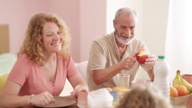 Neşeli çok nesilli aile birlikte sağlıklı bir kahvaltı yapıyorlar. Yaşlı bir adam masada taze bir kırmızı elma soyarken gülümsüyor.