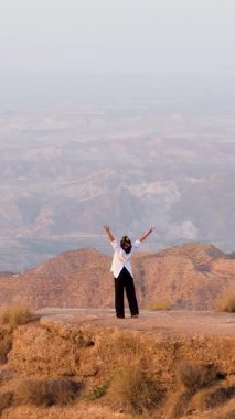 Uçurumun kenarında duran genç bir kadın çölde kollarını neşeyle kaldırıyor. Başarı, özgürlük ve mutluluk hissi.