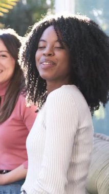 Neşeli, genç, çok ırklı kız arkadaşlar kanepede oturmuş ellerinde içkilerle güneşli bir yaz haftasonu boyunca mutlu bir şekilde kameraya bakıyorlar.