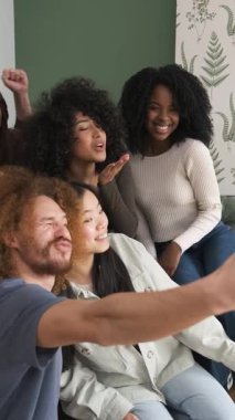 Neşeli, çoklu etnik çeşitliliğe sahip genç erkek ve kadın arkadaşlar, hafta sonu evde akıllı telefon kullanarak selfie çekiyorlar.