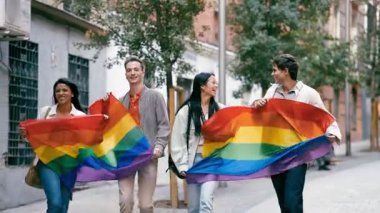 Çok ırklı bir grup arkadaş ellerinde büyük, renkli bir lgbtq gökkuşağı bayrağı tutarken gülerek ve kutlama yaparak şehir caddesinde yürüyorlar.
