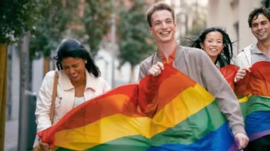 Bir grup mutlu çok kültürlü arkadaş eşcinsellerin geçit töreninde gülümseyip yürüyorlar. Büyük bir gökkuşağı bayrağını bir arada tutuyorlar.