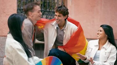 Bir grup genç arkadaş dışarıda gülüp sohbet ediyorlar. Gurur gününü kutlamak için gökkuşağına sarılmış bir adam.