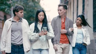Dört genç çok kültürlü arkadaş birlikte yürüyüş yapıyorlar. Asyalı bir kadın yol tarifi için telefonunu kullanırken arkadaşları konuşup etrafa bakınıyor.