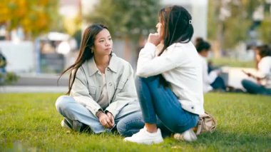 İki Asyalı genç bayan arkadaş, halka açık bir parkta çimlerin üzerinde oturmuş, birlikte geçirecekleri günün tadını çıkarırken ciddi bir konuşma yapıyorlar.