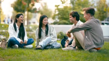 Neşeli çoklu etnik gruplardan oluşan bir arkadaş grubu çimlerin üzerinde oturmuş, keyifli bir sohbetin tadını çıkarıyor ve bir şehir parkında birlikte kahkaha atıyorlar.