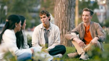 Çimlerin üzerinde yan yana otururken sohbet eden bir grup genç arkadaş. Erkekler ve kadınlar bir şehir parkında dinleniyor ve kaynaşıyorlar.