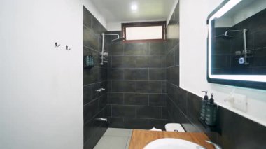 İçinde koyu fayanslar olan sofistike bir banyo, içeri giren yağmur duşu, ve aydınlatılmış aynalı ahşap bir tezgah.