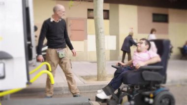 Sosyal hizmet görevlisi engelli bir adama yardım ediyor. Tekerlekli sandalye girişi için özel bir minibüs kullanıyor. Ulaşılabilir ulaşımı gösteriyor.
