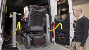 Hidrolik asansörü uzaktan kumandayla çalıştıran adam, elektrikli tekerlekli sandalyedeki birinin ulaşılabilir bir minibüse binmesine yardım ediyor.