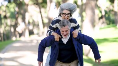 Mutlu olgun koca, karısını güneşli bir parkta sırtında taşıyor. Kaygısız ve sevgi dolu son sınıf çifti birlikte neşeli bir anın tadını çıkarıyor.